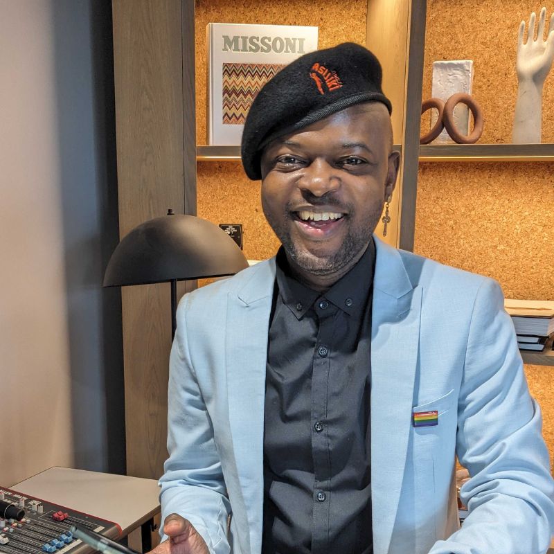 Ethan - SafePlace Internation A person in a light blue blazer and black beret smiles indoors, standing by a desk with shelves, books, and decor items in the background. SafePlace International
