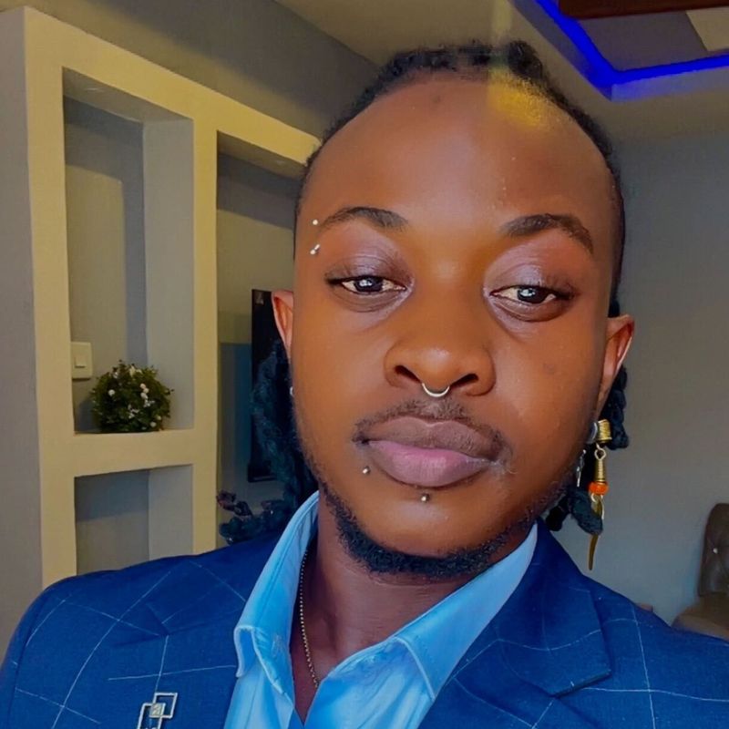 Francis - SafePlace Internation A person with facial piercings and braided hair wears a blue suit and light blue shirt, posing indoors with shelves and a small plant in the background. SafePlace International