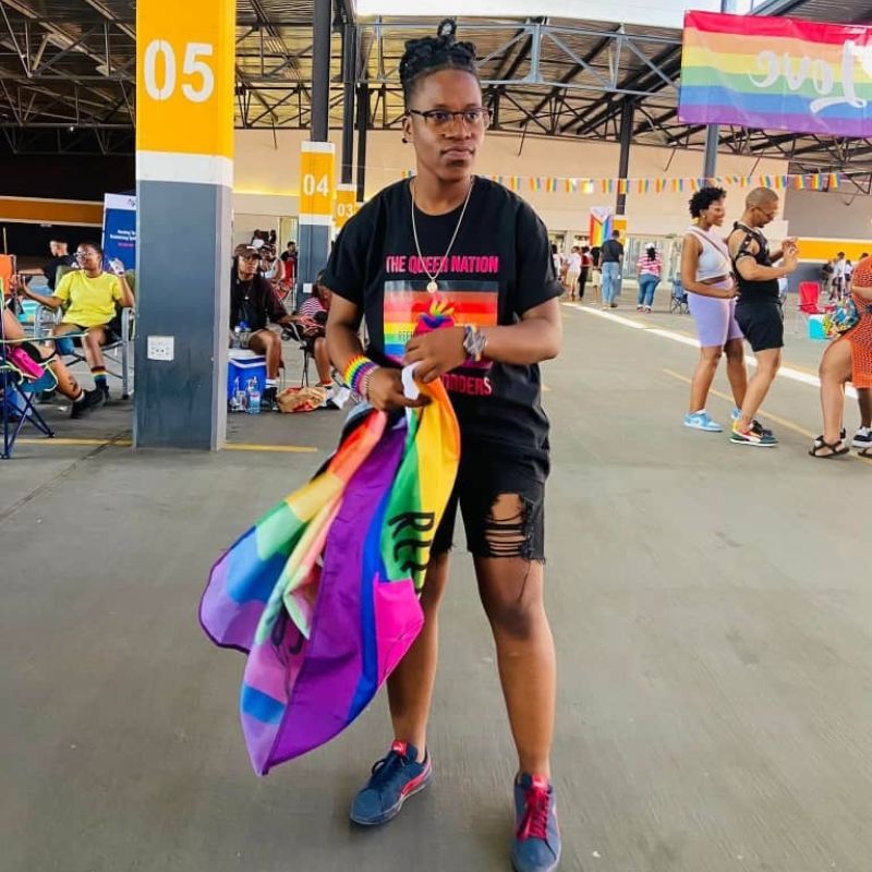 GG - SafePlace Internation Person holding a rainbow flag stands in an indoor space with other people; wearing a black T-shirt with a rainbow design and ripped black shorts. Pride banners are visible in the background. SafePlace International
