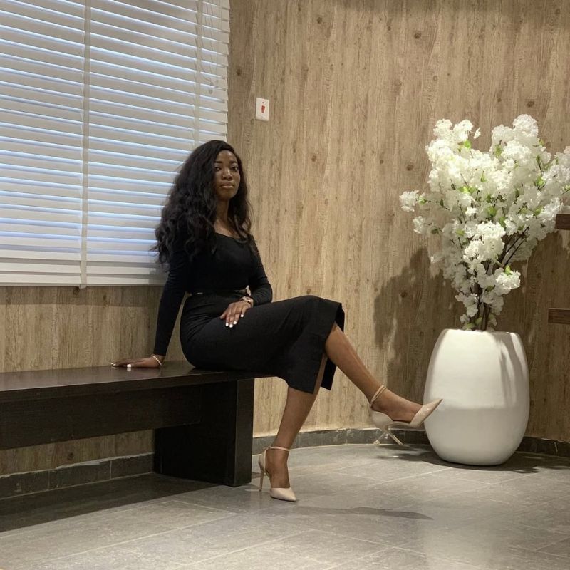 Mercy - SafePlace Internation Woman in a black dress sits on a wooden bench near a large white vase with white flowers, next to a window with closed blinds in a modern room. SafePlace International