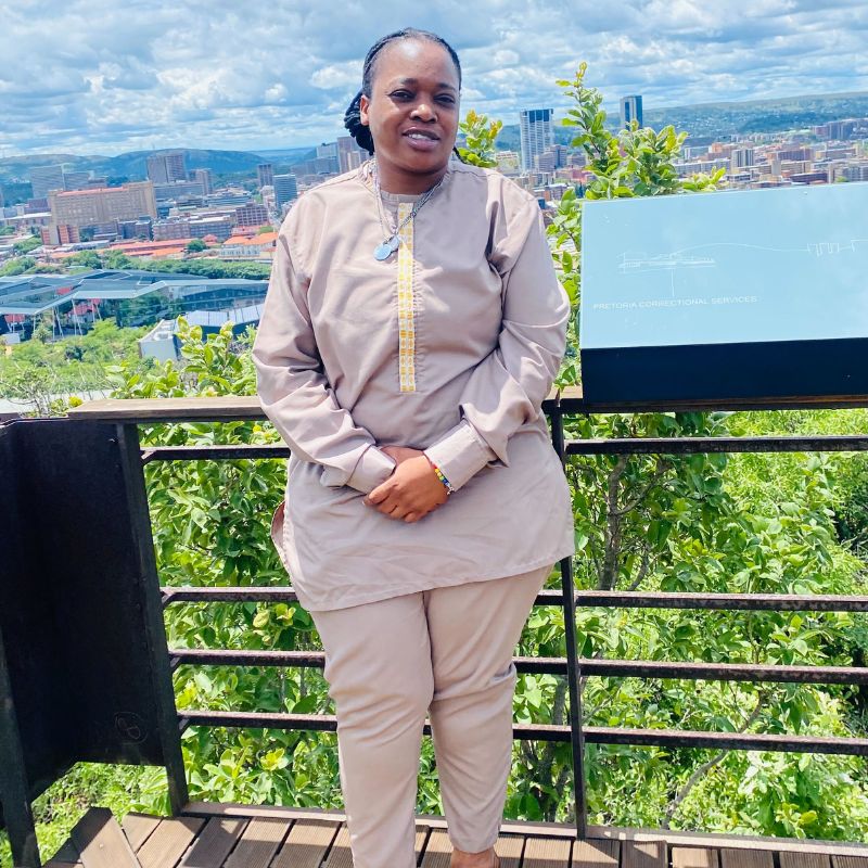 Nyasha “Masi” Zhakata - SafePlace Internation A person in beige clothing stands by a railing overlooking a cityscape with greenery and buildings under a partly cloudy sky. SafePlace International