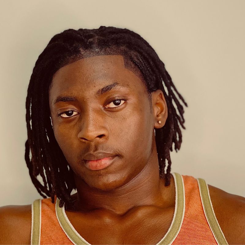 Chad Semu - SafePlace Internation A person with medium-length dreadlocks, wearing an orange tank top with green trim, poses in front of a plain beige background, representing the SafePlace International Team. SafePlace International