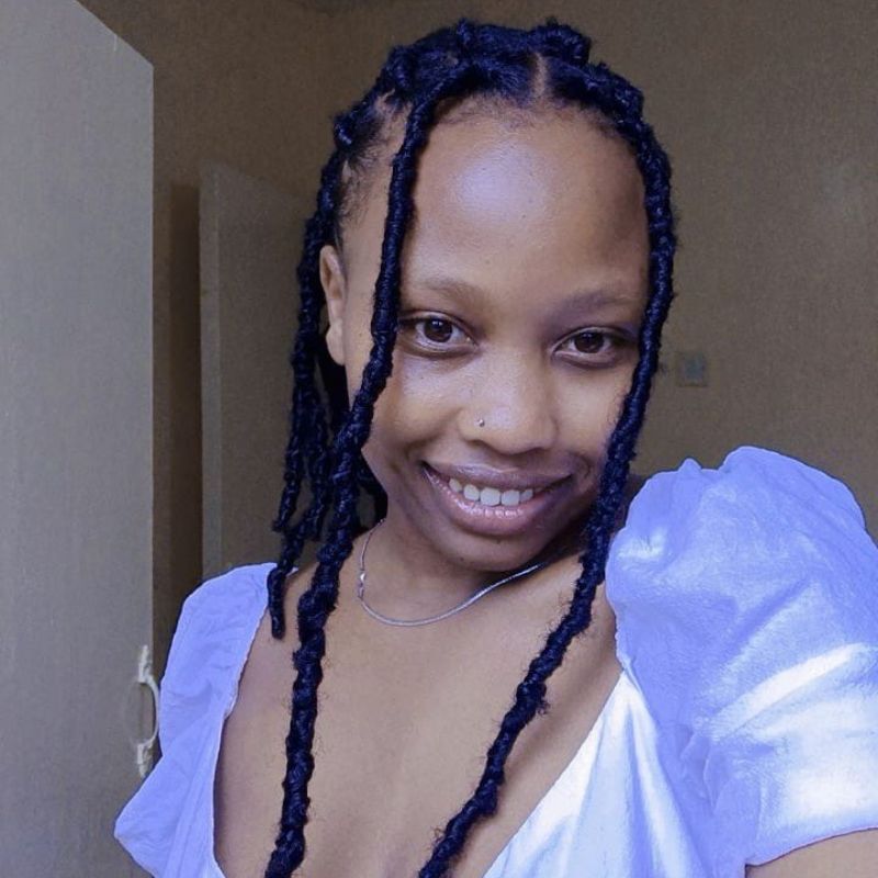 JESS WANGARI - SafePlace Internation A person with braided hair smiles at the camera, wearing a white puff-sleeve top and a thin necklace, standing indoors with a door in the background. SafePlace International